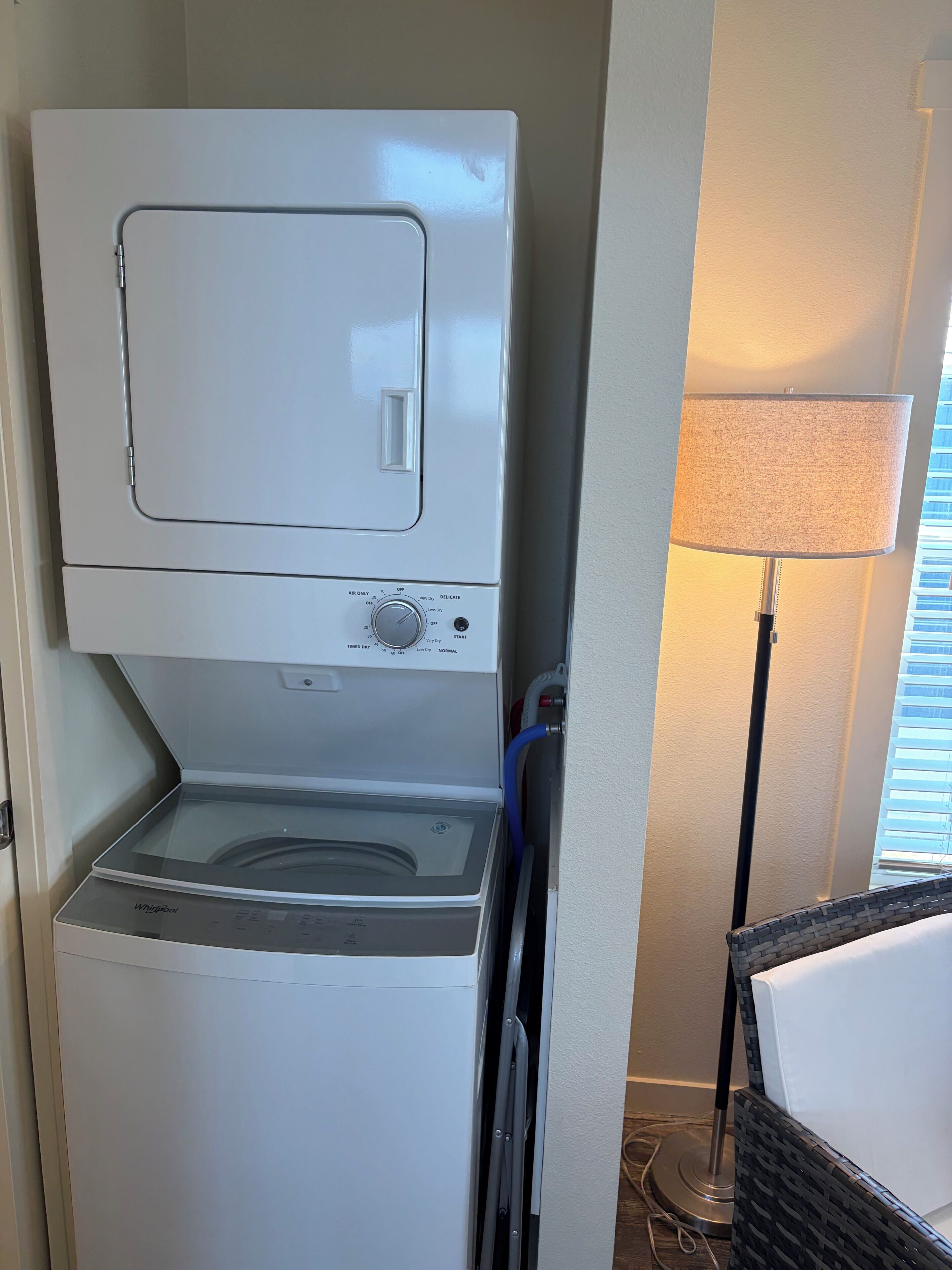 En kombineret vaskemaskine og tørretumbler med kontrolpanel, placeret i et hjørne af et rum med en lampe og en fletstol.