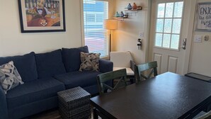 Una sala de estar con un sofá azul oscuro, una pequeña mesa de madera y una silla. Hay un cuadro en la pared y una ventana con persianas.