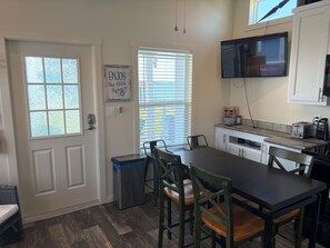 A compact kitchen with a dining table, chairs, a trash bin, a television mounted on the wall, and a window with blinds. - Lakefront Tiny Home with Dock & View (Kerens)