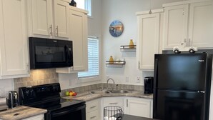 Una cocina moderna con armarios blancos, un refrigerador negro, un microondas y una estufa. Hay una mesa de comedor con sillas y un ventilador de techo.
