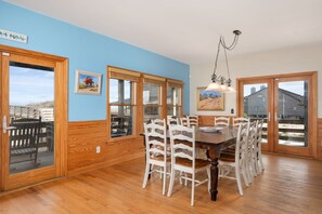 Une salle à manger avec une table et des chaises en bois, de grandes fenêtres et une vue sur l’extérieur.