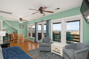 Une pièce dotée d’un ventilateur de plafond, d’un coin repas avec des chaises et une table en bois, d’un fauteuil gris, d’un tapis bleu et d’une vue sur la plage, visible à travers de grandes fenêtres.