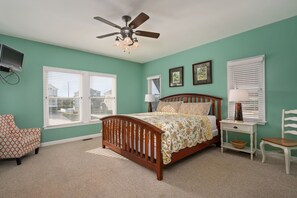 Une chambre à coucher comprenant un lit, un ventilateur de plafond, un téléviseur fixé au mur et deux fenêtres équipées de stores.