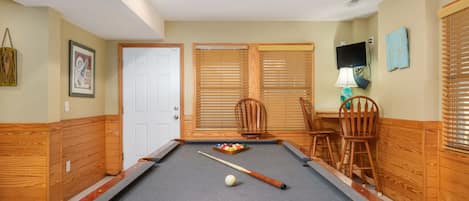 A pool table with a cue stick, a ball, and a set of billiard balls. There are wooden chairs and a wooden bench.