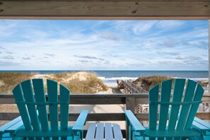 En træterrasse med to turkise Adirondack-stole og et træbord, med udsigt over en strand med klitter og havet.