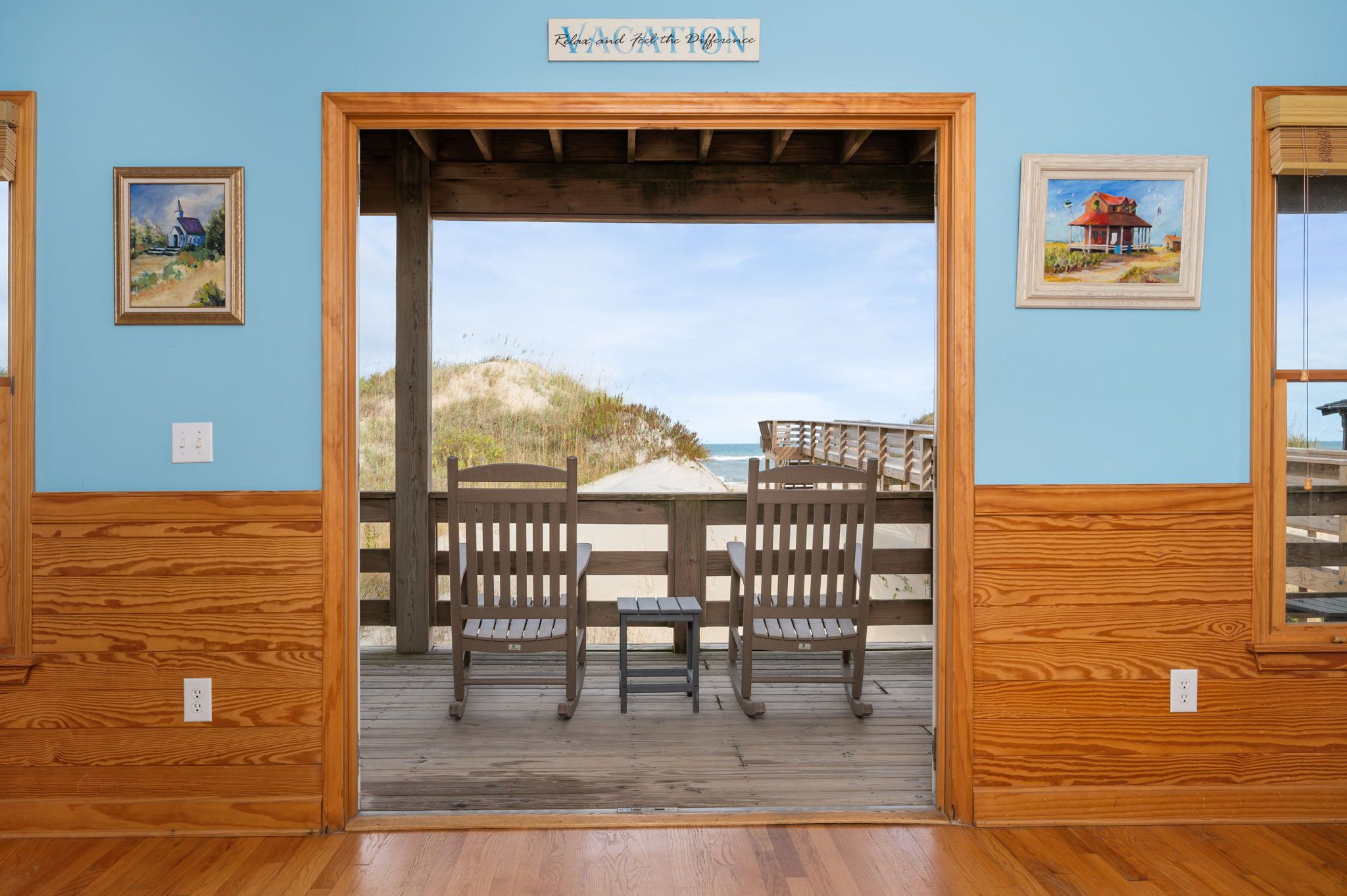 A wooden deck with two chairs and a table, overlooking a beach with dunes and a clear sky.