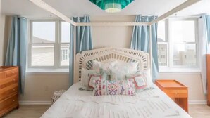 Une chambre avec un lit blanc, un ventilateur de plafond et une vue sur les immeubles par les fenêtres.