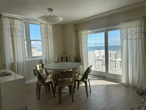 A dining area with a round table and chairs, large windows with curtains, and a view of the ocean.