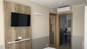 Moderni hotellihuone, jossa on puukoristeinen seinä, seinälle asennettu televisio, pieni hylly, valkoisilla tekstiileillä varustettu sänky ja näkymä kylpyhuoneeseen.