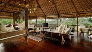 A spacious, open-air lounge with wooden flooring and a thatched roof, featuring a sofa, a coffee table, and a television. - Villa Inka Ubud by Nakula (Ubud)