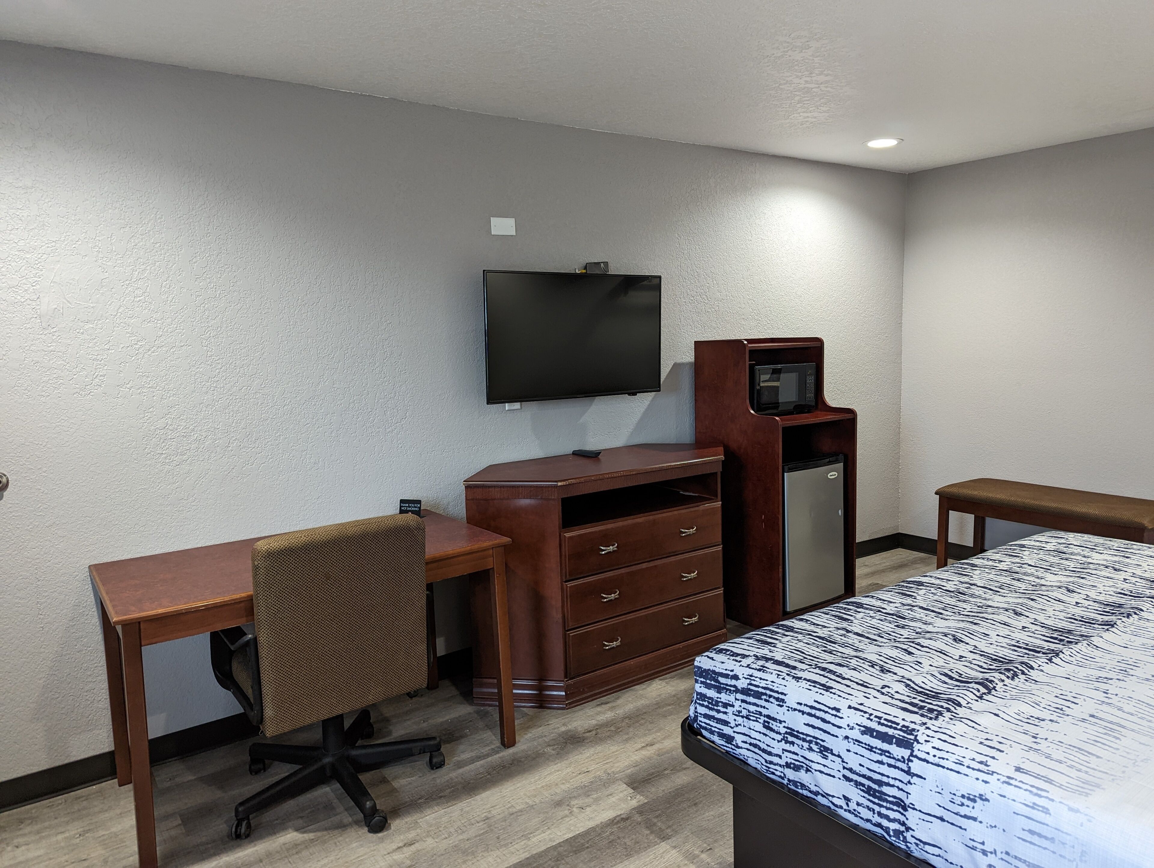 A hotel room with a wooden desk, a chair, a bed with a patterned bedspread, a television mounted on the wall, and a small refrigerator.