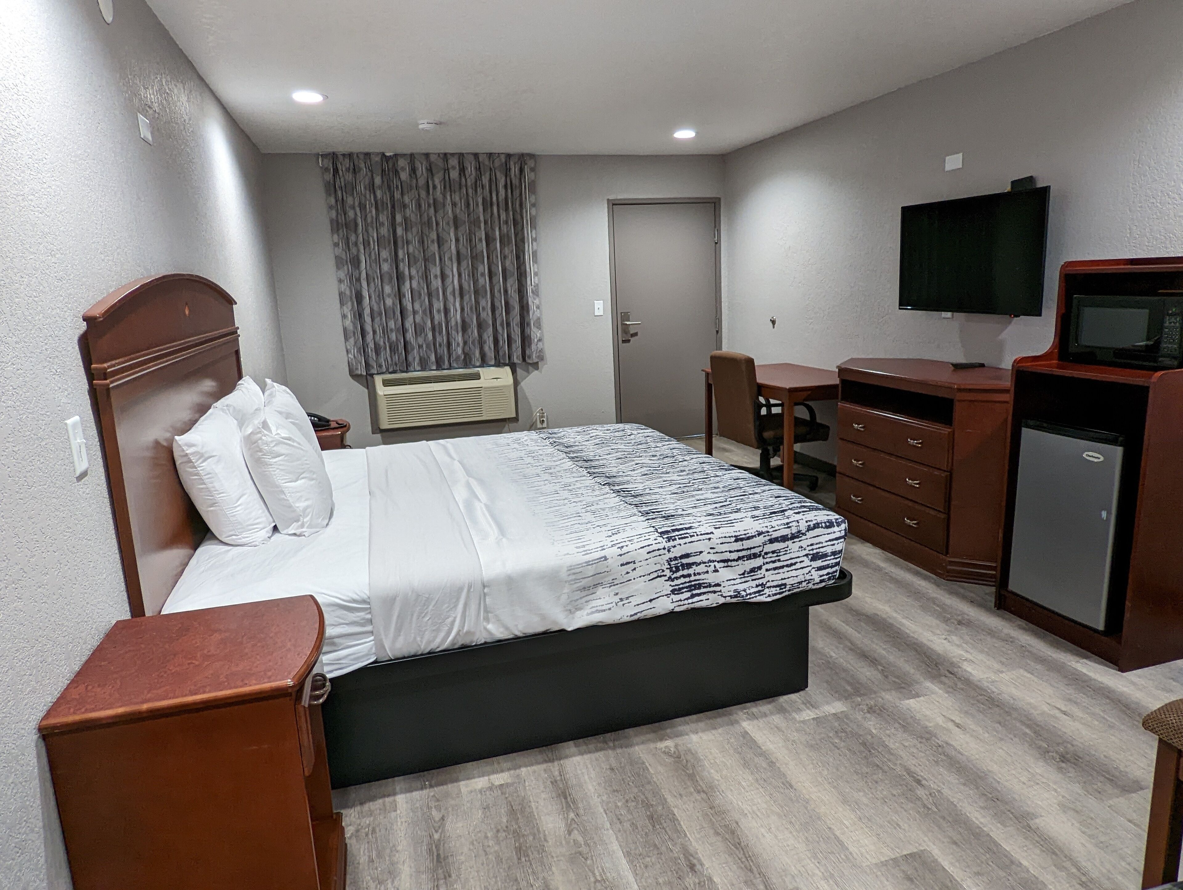 A hotel room with a bed, a desk, a chair, a TV, a wardrobe, and a small refrigerator.