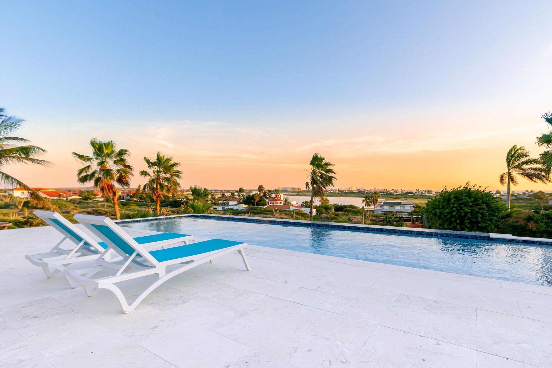 Une piscine entourée de chaises longues, bordée de palmiers et offrant une vue sur le paysage urbain au coucher du soleil.