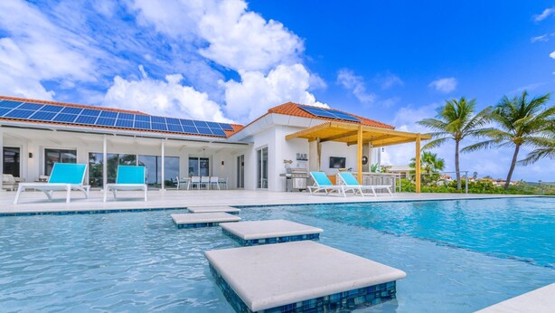 A modern pool area with a clear blue sky, a pool with floating steps, and lounge chairs. - Caya di Bista Bunita 6 (Noord)