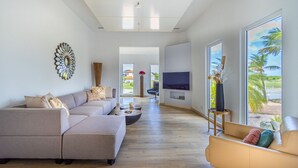 A spacious living room with a sofa, coffee table, and TV, large windows with a view of palm trees. - Caya di Bista Bunita 6 (Noord)