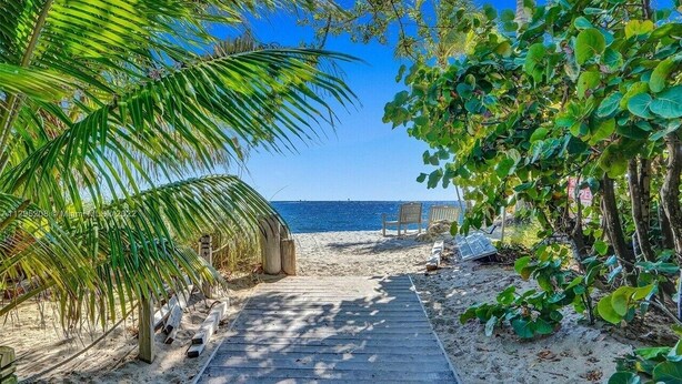 A wooden pathway leading to a beach with a chair and a table, surrounded by lush greenery.