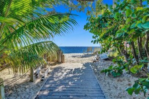 Ein Holzweg führt zu einem Strand, wo ein Stuhl und ein Tisch inmitten üppiger Vegetation stehen.