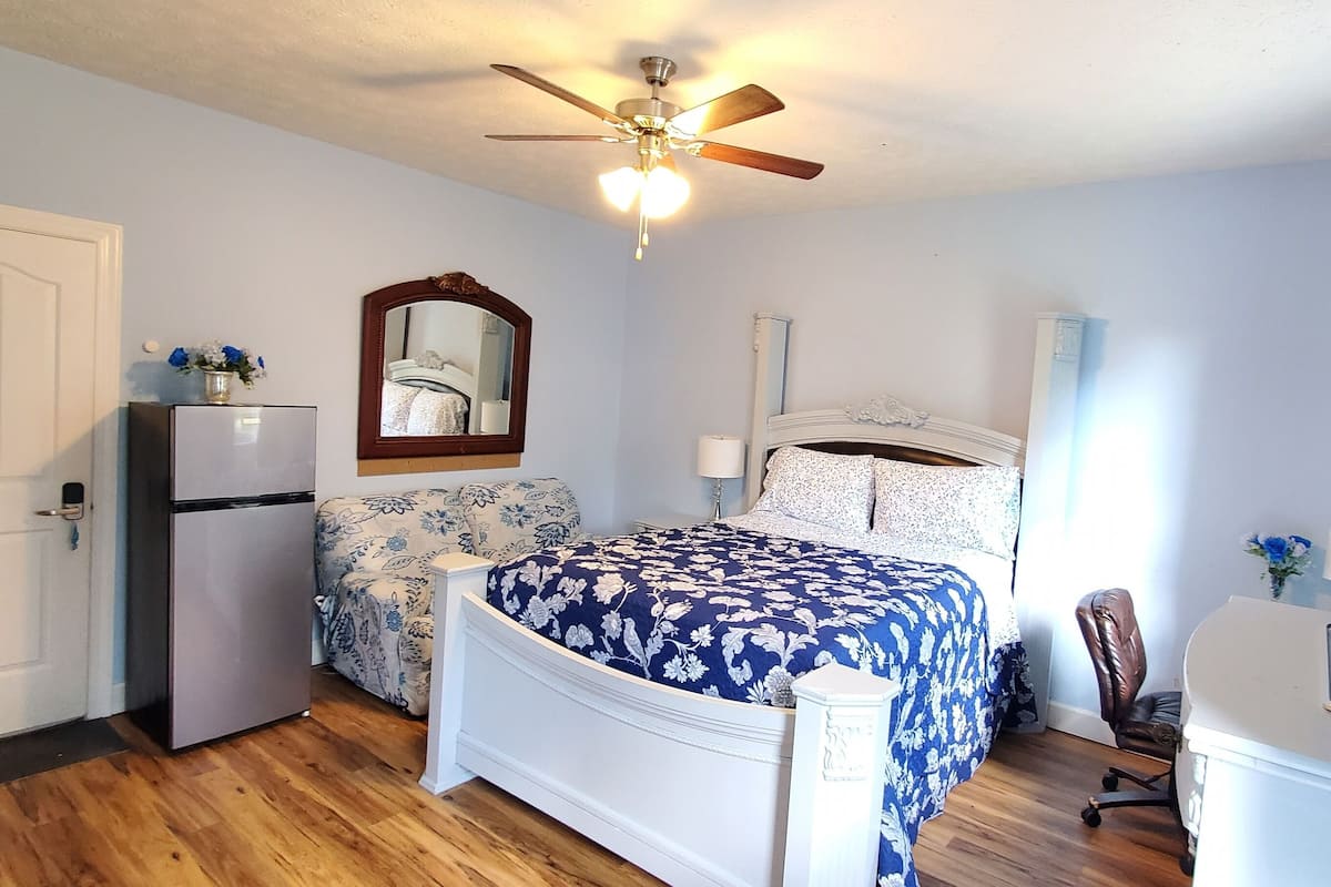 Un dormitorio con cama, ventilador de techo, refrigerador y escritorio con silla.