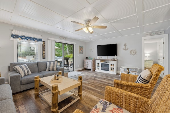 A living room with a sofa, a wooden coffee table, wicker chairs, a ceiling fan, a television, and a kitchen area with cabinets and a countertop.