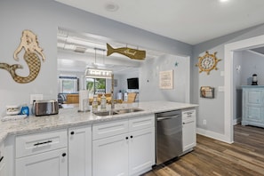 A kitchen with white cabinets, a stainless steel dishwasher, a marble countertop, and decorative wall art including a mermaid and a fish sign.