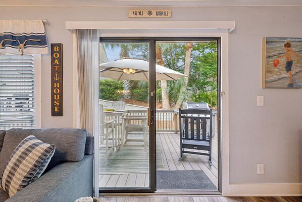 Una sala de estar con un sofá, un letrero que dice 'BOATHOUSE' y un ventanal corredizo que da a una terraza exterior con muebles de patio y una parrilla.