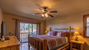 Un lit en bois avec une tête de lit, un ventilateur de plafond, une table de chevet avec une lampe et une fenêtre avec des rideaux.