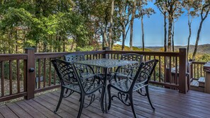 Une terrasse en bois avec une table et des chaises en métal, entourée d’arbres et offrant une vue sur des collines lointaines.