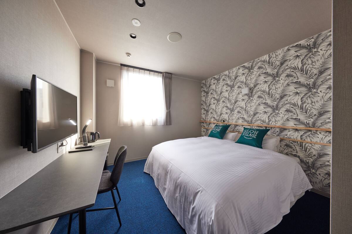 A hotel room with a bed, a desk with a flat-screen TV, a chair, and a window with curtains.