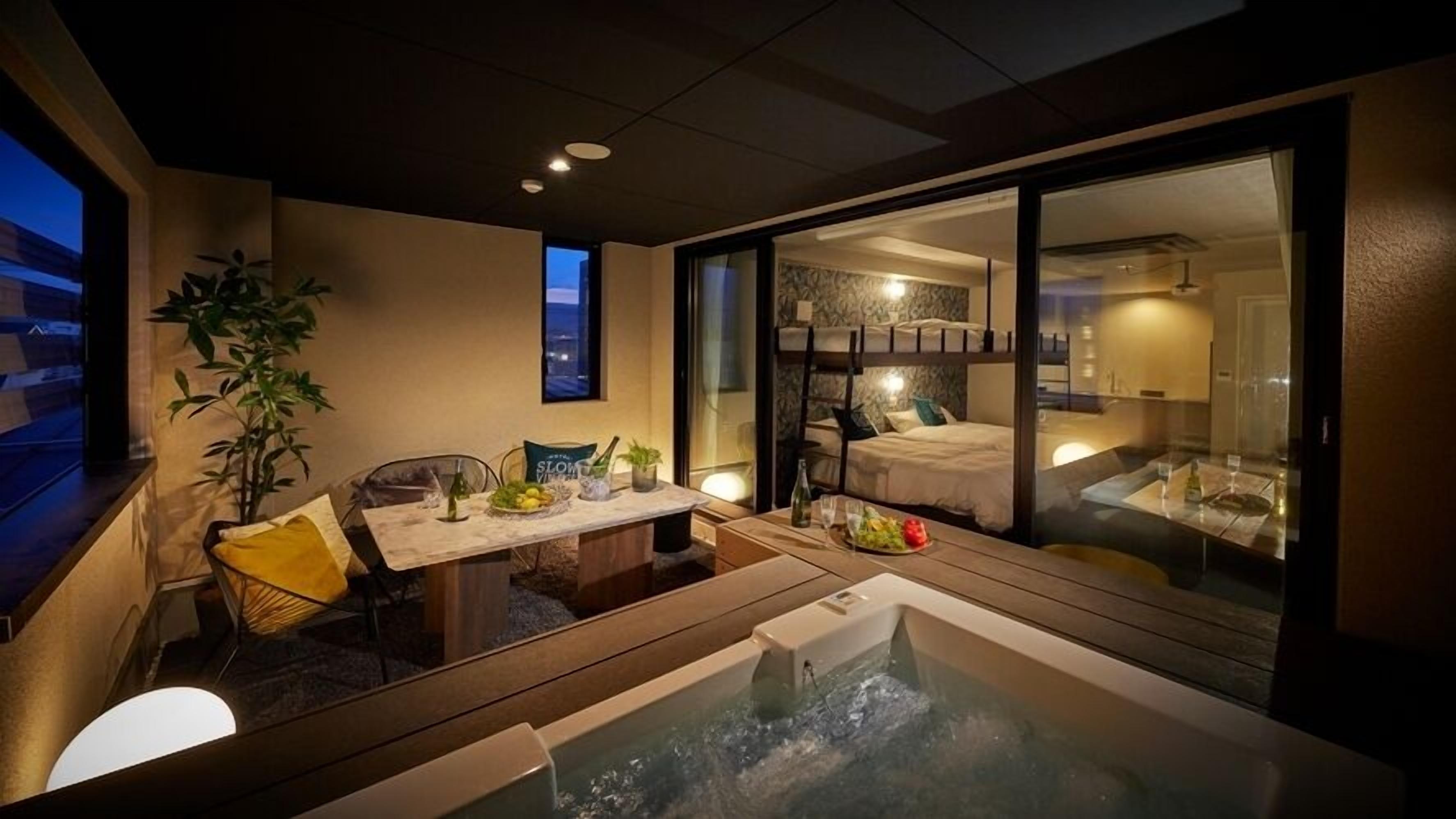 A modern hotel room with a glass-enclosed balcony, a dining area with a table set for two, and a hot tub.