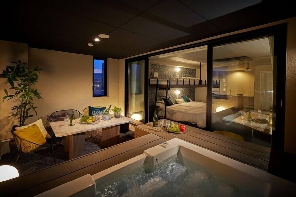 A modern hotel room with a glass-enclosed balcony, a dining area with a table set for two, and a hot tub.