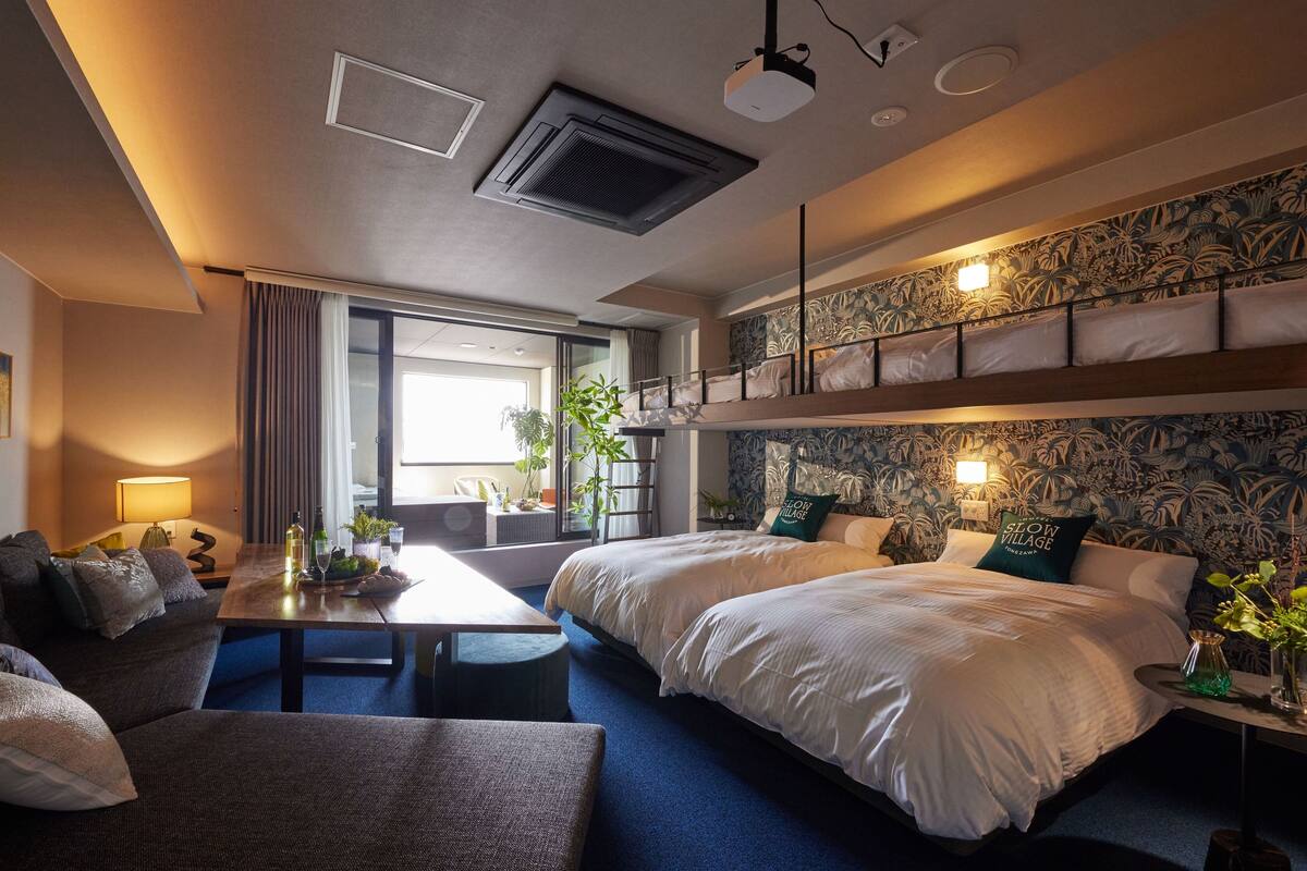 A hotel room with a large bed, a sofa, a dining table with a lamp, and a balcony with a view.