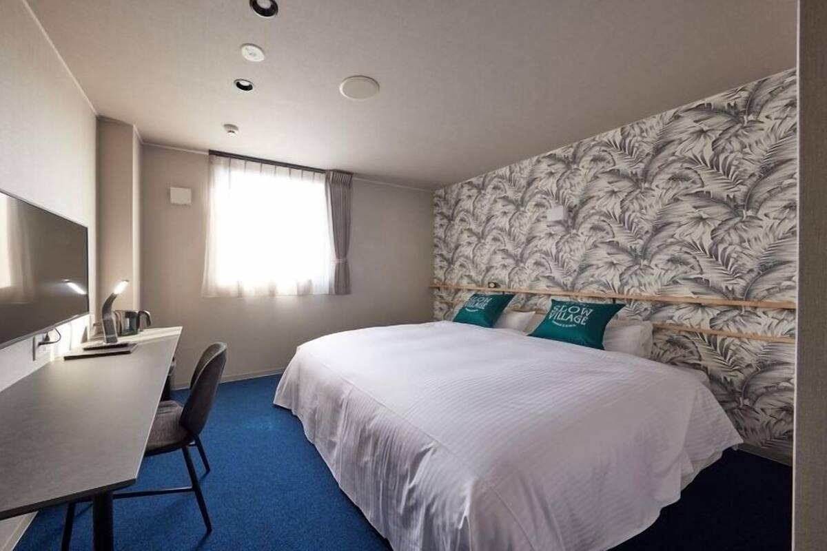 A hotel room with a bed, a desk with a TV, a chair, and a window with curtains.