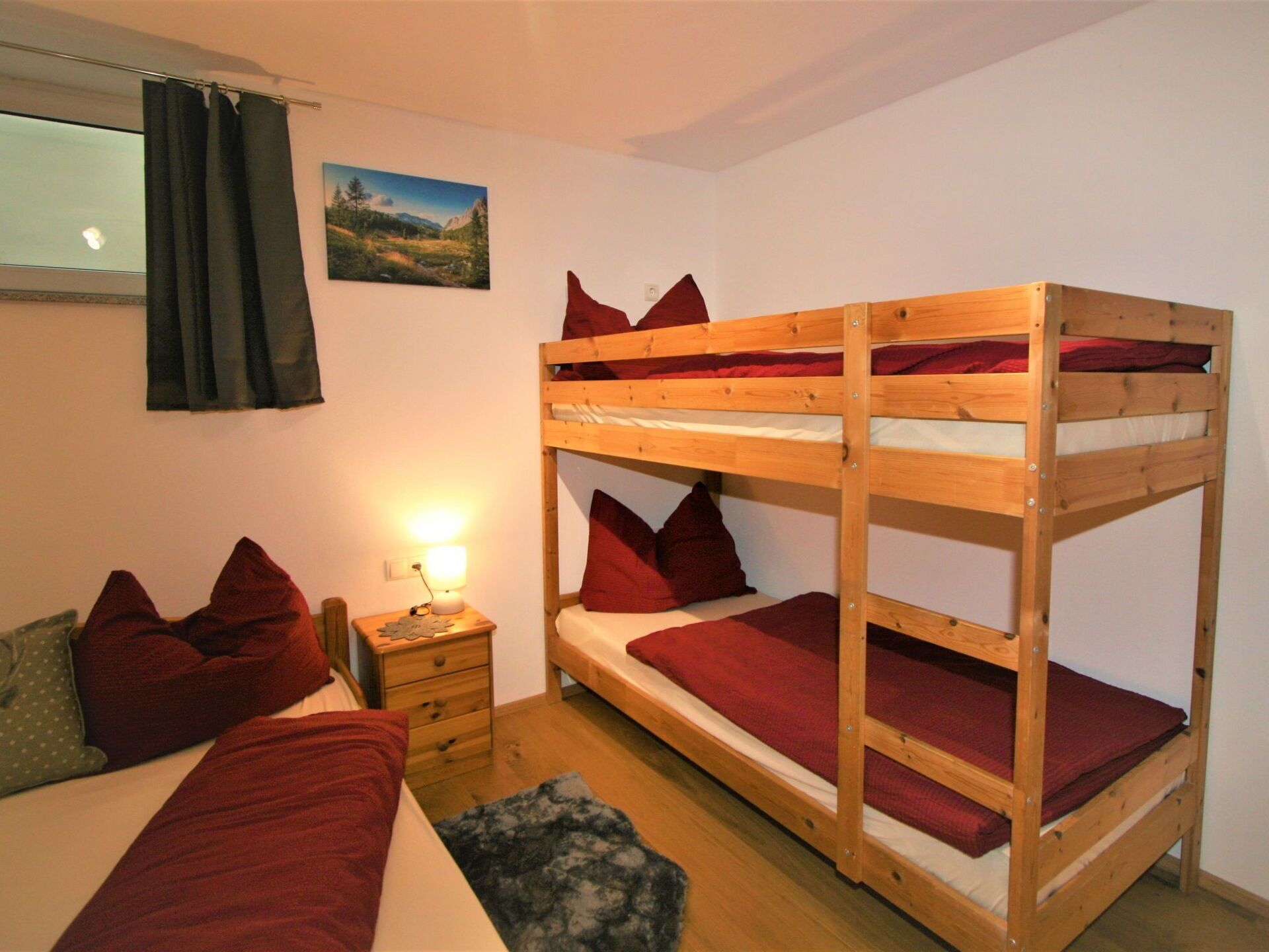 Une chambre avec un lit superposé en bois, une table de chevet avec une lampe, un petit tiroir et une fenêtre avec des rideaux.