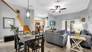 A modern living room with a gray sofa, a dining table with chairs, a kitchen area, and a ceiling fan.
