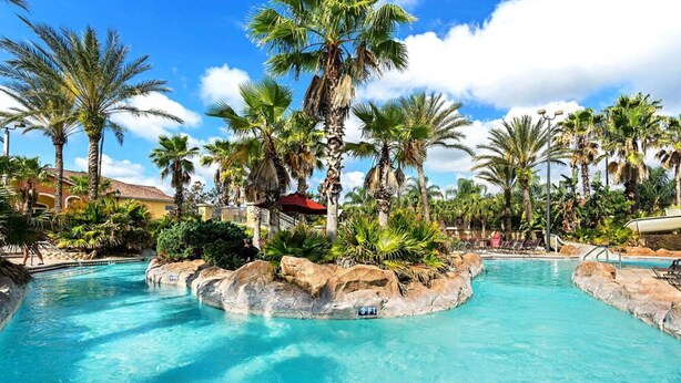 A pool area with palm trees and a red umbrella, surrounded by rocks and greenery.