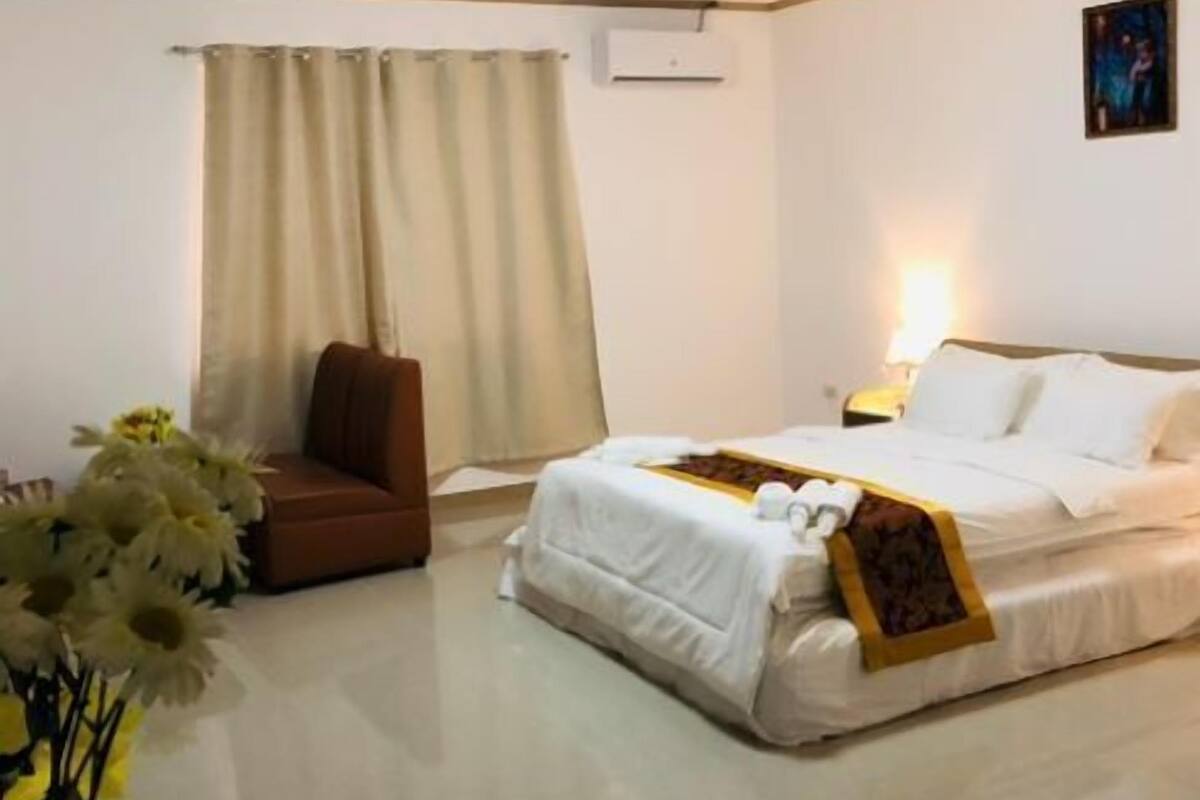 A hotel room with a bed, a brown armchair, a small table with a vase of flowers, and a painting on the wall.
