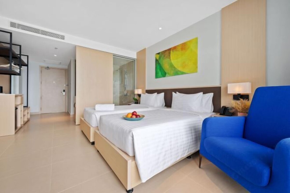 A modern hotel room with a large bed, a blue armchair, a painting on the wall, and a small table with a fruit bowl.