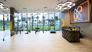 A spacious gym with modern equipment, large windows offering views of palm trees, and a neon sign indicating the name 'Aqua Gym'. - The Empyrean Cam Ranh Beach Resort (Bac Cam Ranh)