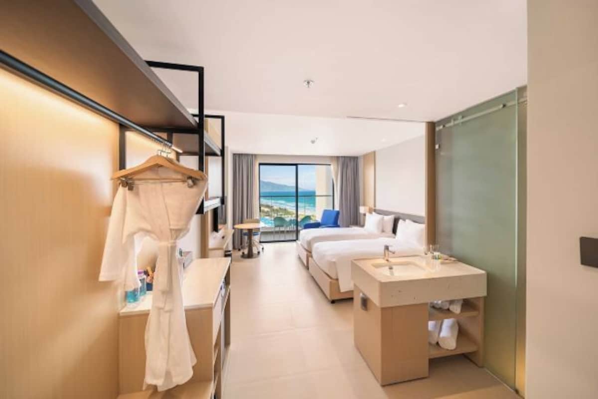 A modern hotel room with a bed, a desk, a vanity, and a large window with a view of the sea.