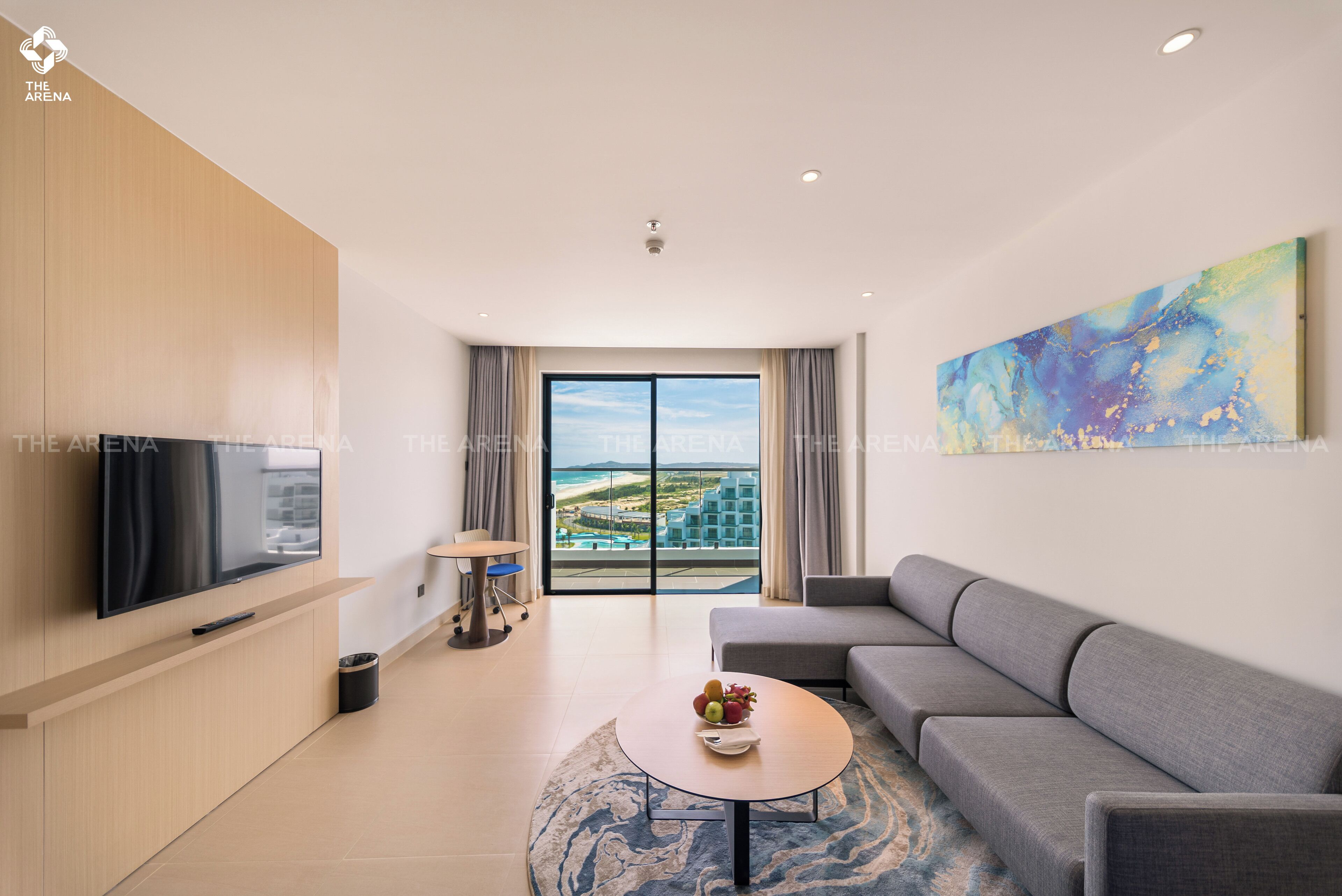 A modern living room with a flat-screen TV, a grey sofa, a coffee table with fruit, and a large window with a view of buildings and a beach.