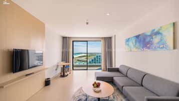 A modern living room with a flat-screen TV, a grey sofa, a coffee table with fruit, and a large window with a view of buildings and a beach.