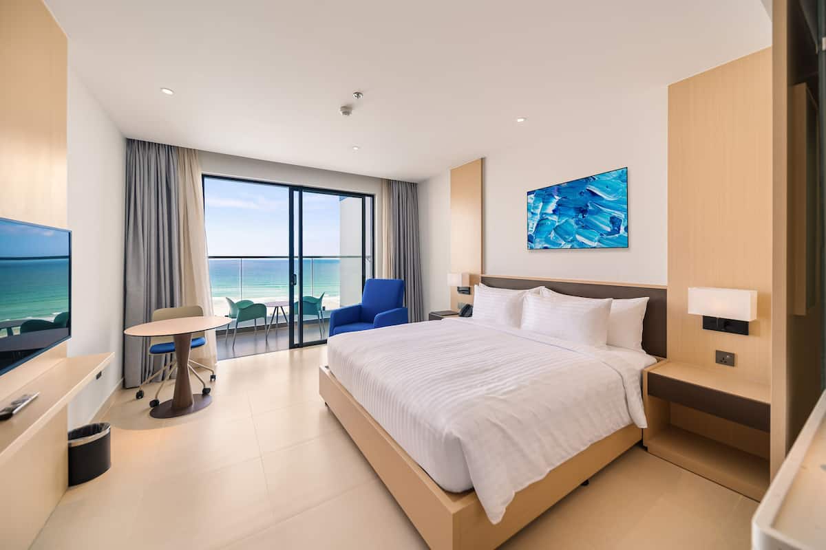 A modern hotel room with a large bed, a desk with a chair, a TV, and a view of the ocean.