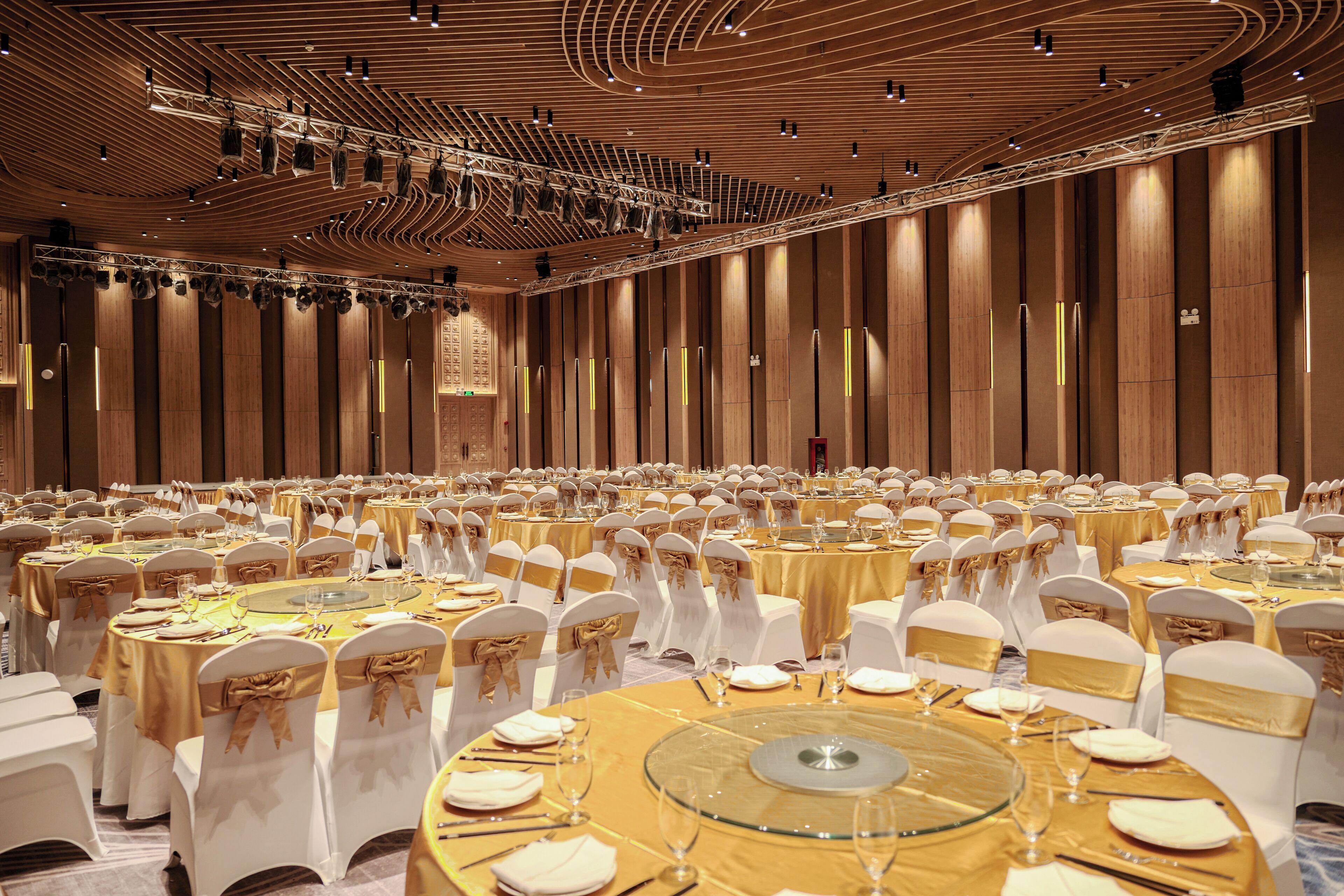 Un amplio salón de banquetes con mesas redondas dispuestas para cenar, sillas blancas con cintas doradas y un techo de madera con lámparas.