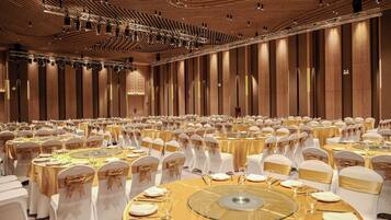Un amplio salón de banquetes con mesas redondas dispuestas para cenar, sillas blancas con cintas doradas y un techo de madera con lámparas.