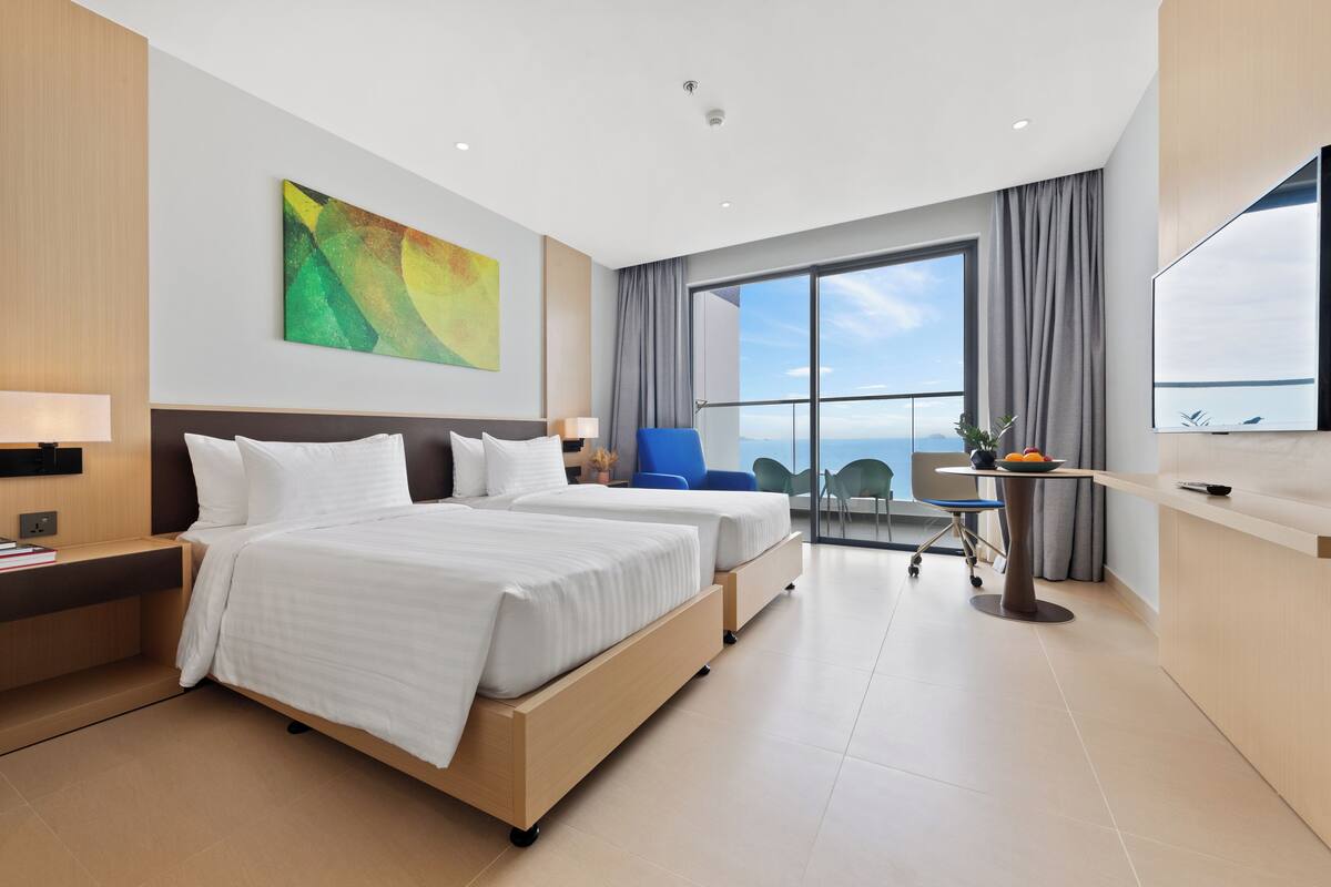 A modern hotel room with two beds, a large window, a flat-screen TV, and a dining area with a table and chairs.