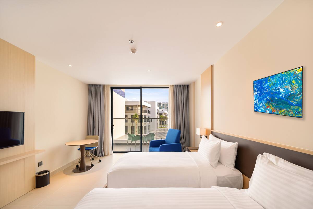 A hotel room with a bed, a desk, a chair, a TV, and a balcony with a view of buildings.