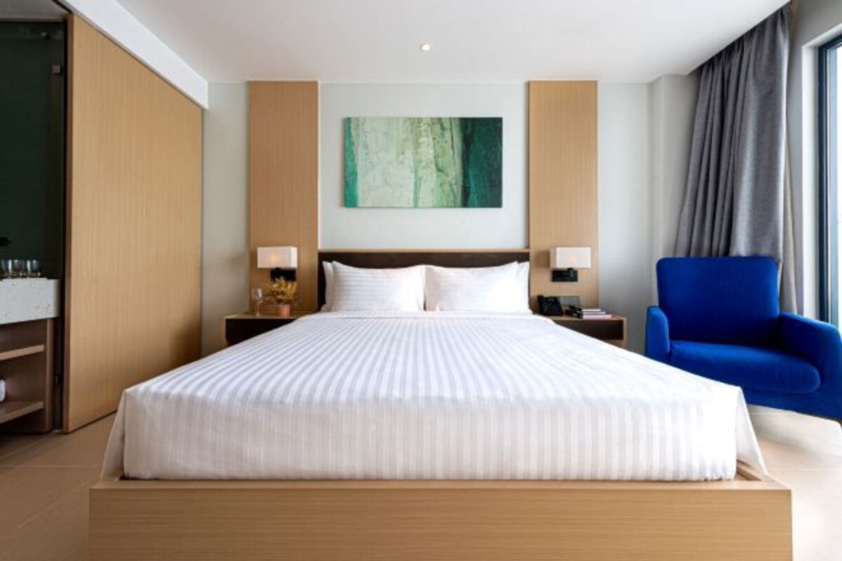 A hotel room with a large bed, two bedside tables with lamps, a blue armchair, and a painting on the wall.