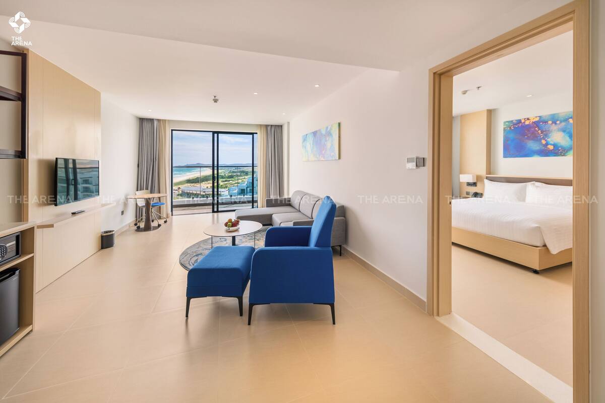 A modern hotel room with a bed, a seating area with a table and chairs, a TV, and a large window with a view of the beach.