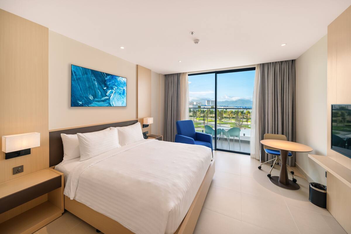 A modern hotel room with a large bed, a desk, a chair, and a view of the city.