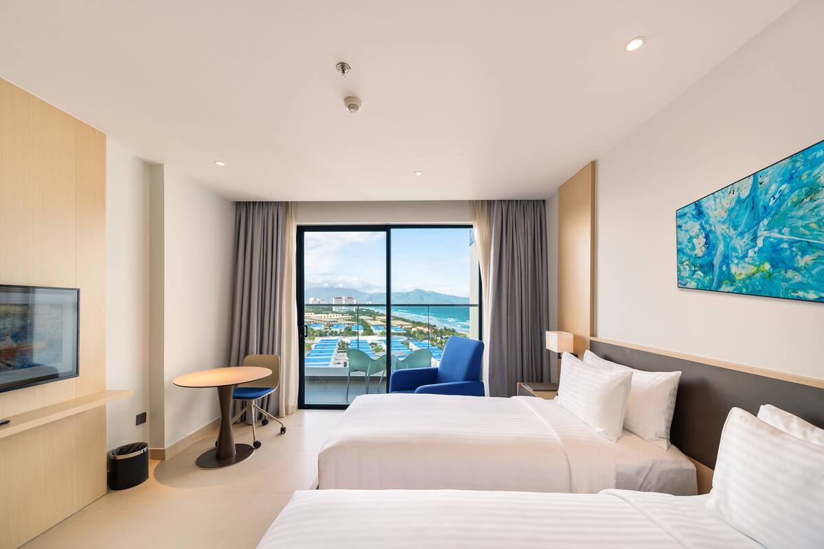 A hotel room with a bed, a desk, a chair, a TV, and a large window with a view of a resort and ocean.